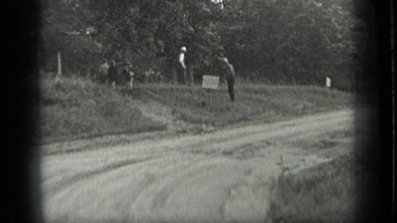 Dirt road beside trees with several distant people near a small sign.