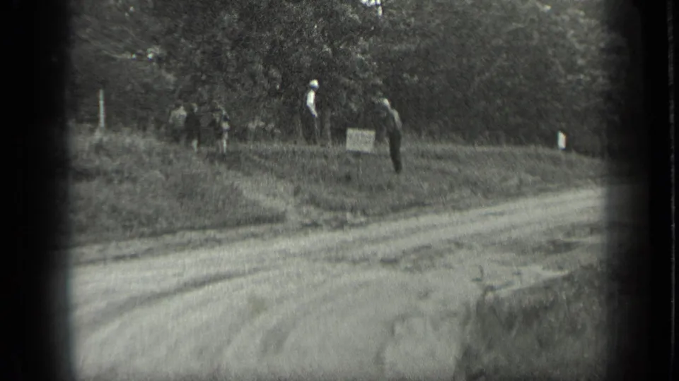 Long Lake, Canada home-movie frame showing a dirt road, tree line, distant figures, and a small roadside sign in 1939 footage.
