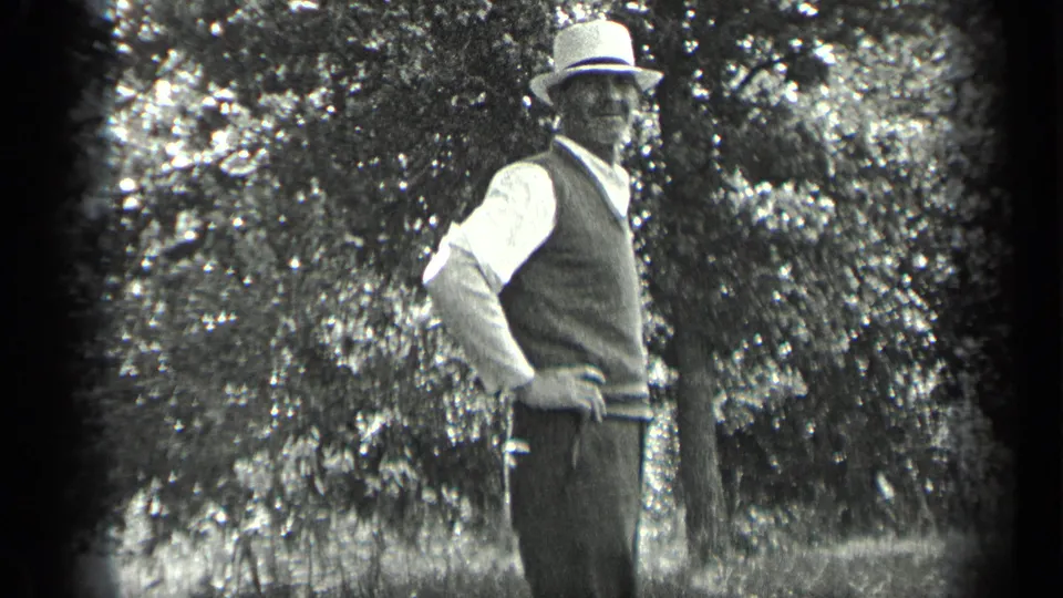 Close home-movie view of the posed man in a light hat, light shirt, and dark vest against trees in Long Lake, Canada.