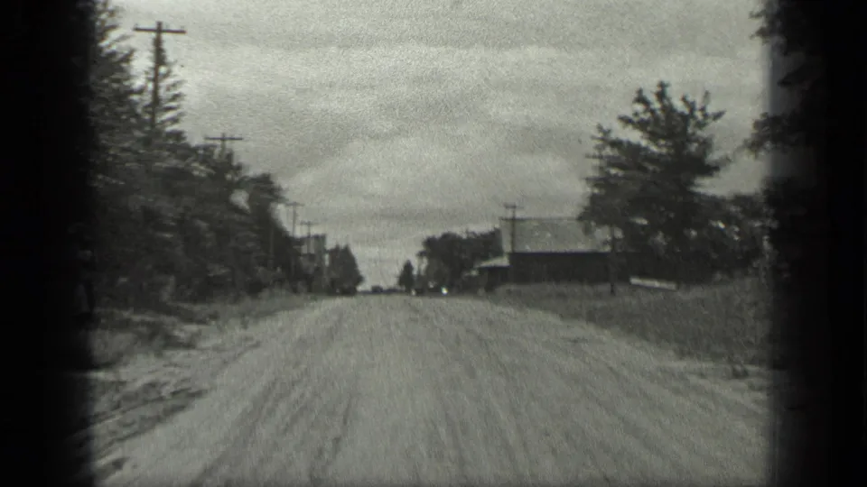 1939 Long Lake, Canada home-movie frame showing a broad unpaved road lined with trees, utility poles, and low roadside buildings.