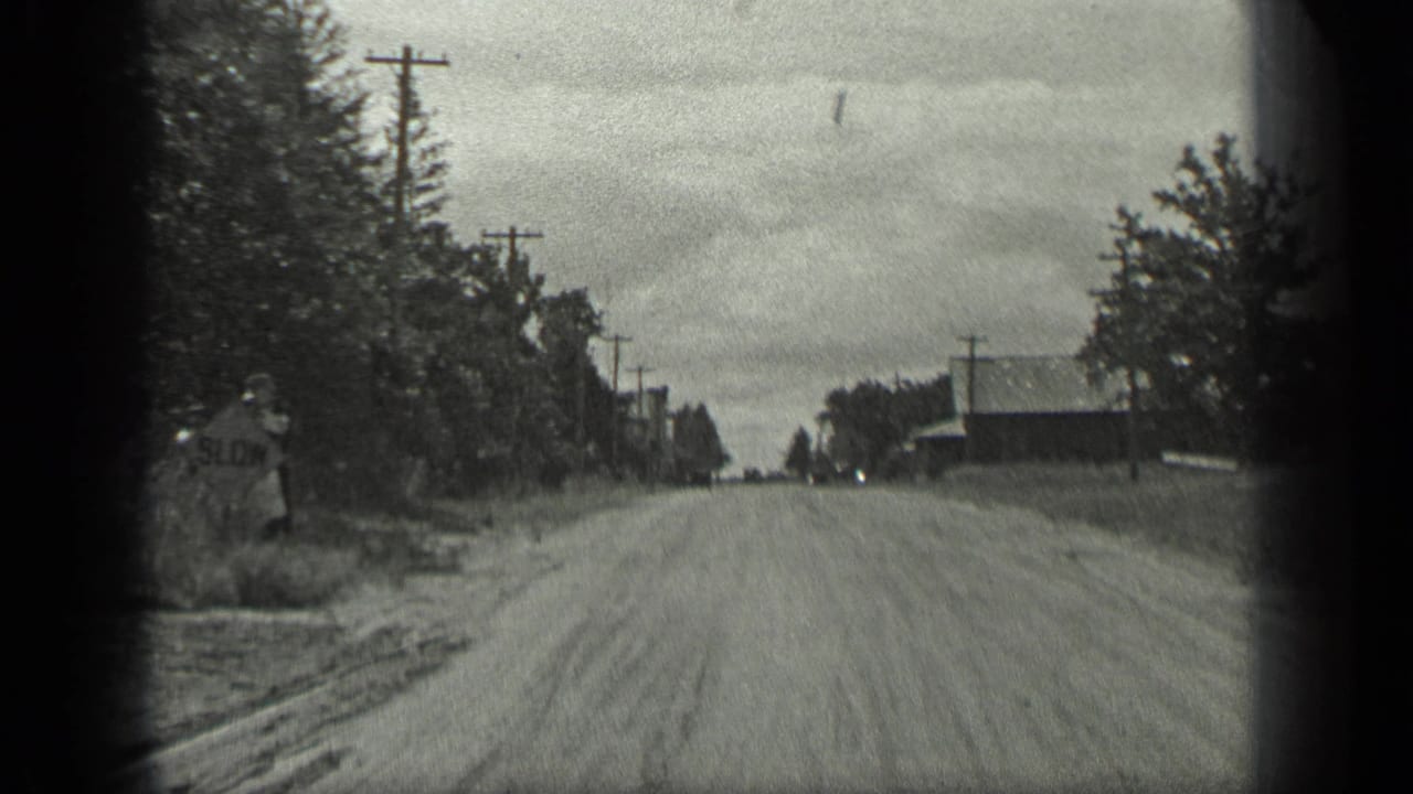 Rural dirt road with a partially visible roadside sign, trees, poles, and buildings.