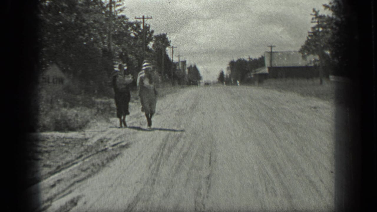 Two women in hats and dresses walk toward the camera on a Long Lake, Canada dirt road, with trees, poles, and buildings nearby.