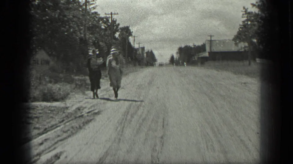 Two women in hats and dresses walk toward the camera on a Long Lake, Canada dirt road, with trees, poles, and buildings nearby.