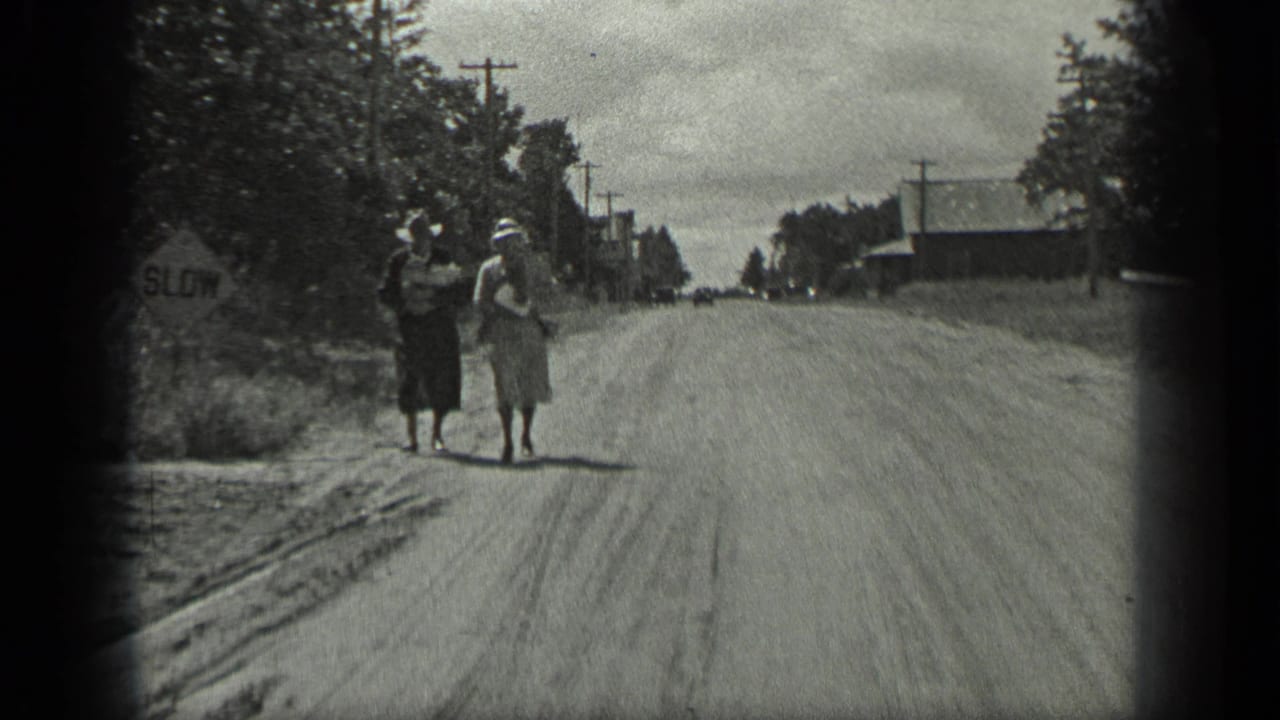 Two women walk along a dirt road carrying packages near a SLOW sign.