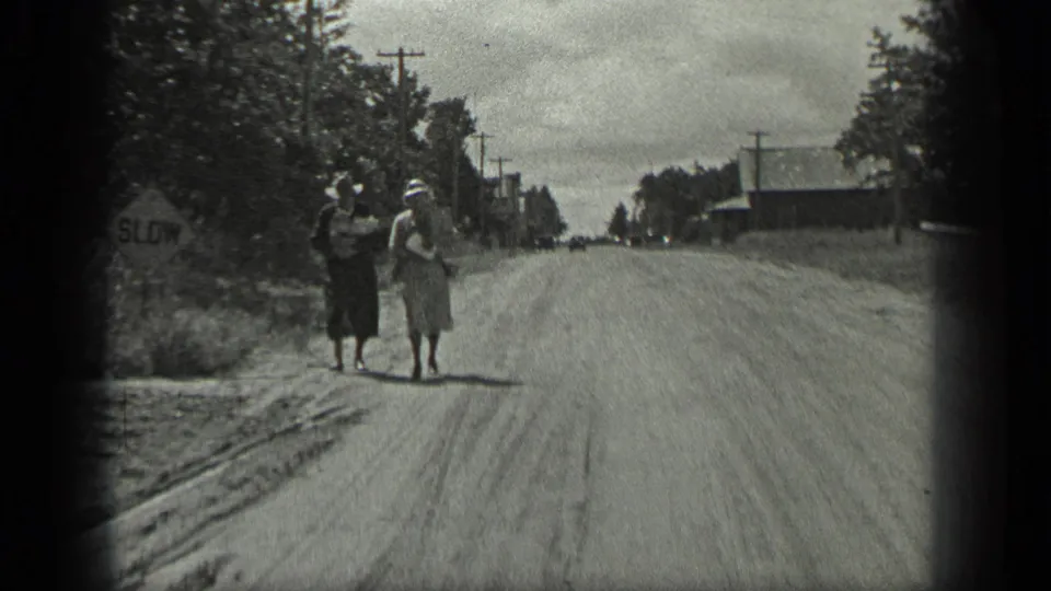 Two women in 1930s dress walk on a dirt road in Long Lake, Canada, with packages, trees, utility poles, and a SLOW sign visible.
