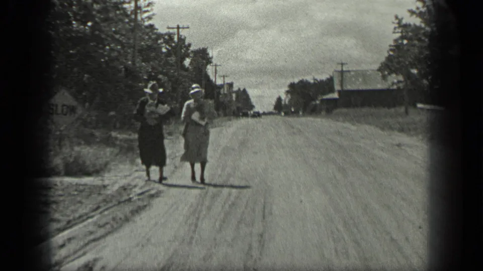 The women continue along the Long Lake dirt road, carrying packages past roadside trees, a SLOW sign, utility poles, and distant buildings.