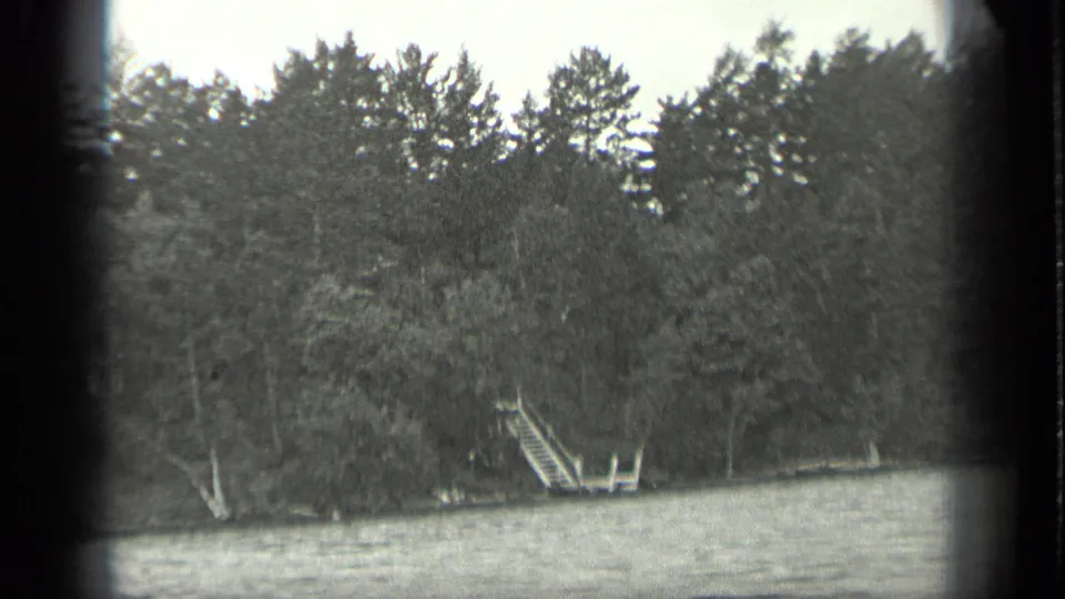 1939 Long Lake, Canada home-movie frame showing rippling water, dense trees, and a small wooden dock with a ladder on the far shore.