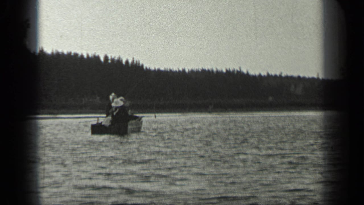 Two people sit in a small boat on a lake near a wooded shoreline.