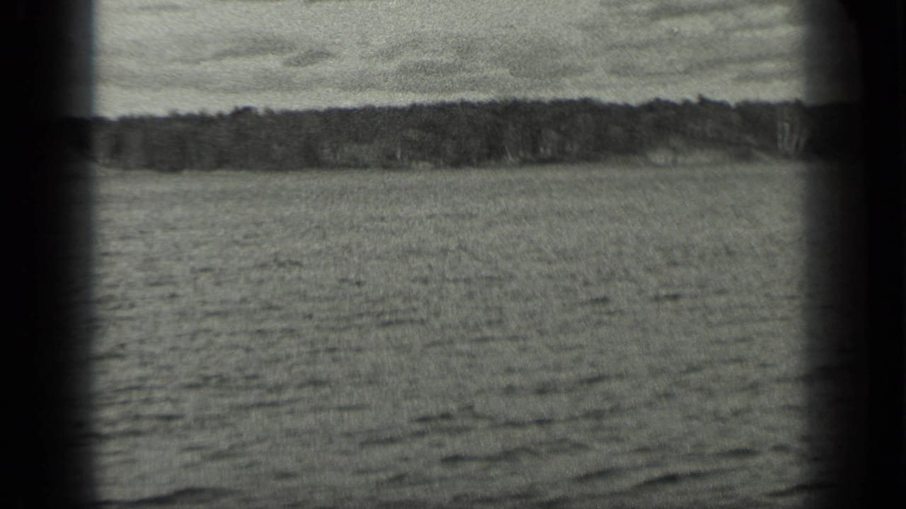 A wide view across Long Lake shows rippled water, a distant tree line, and cloudy sky in black-and-white 8mm footage.