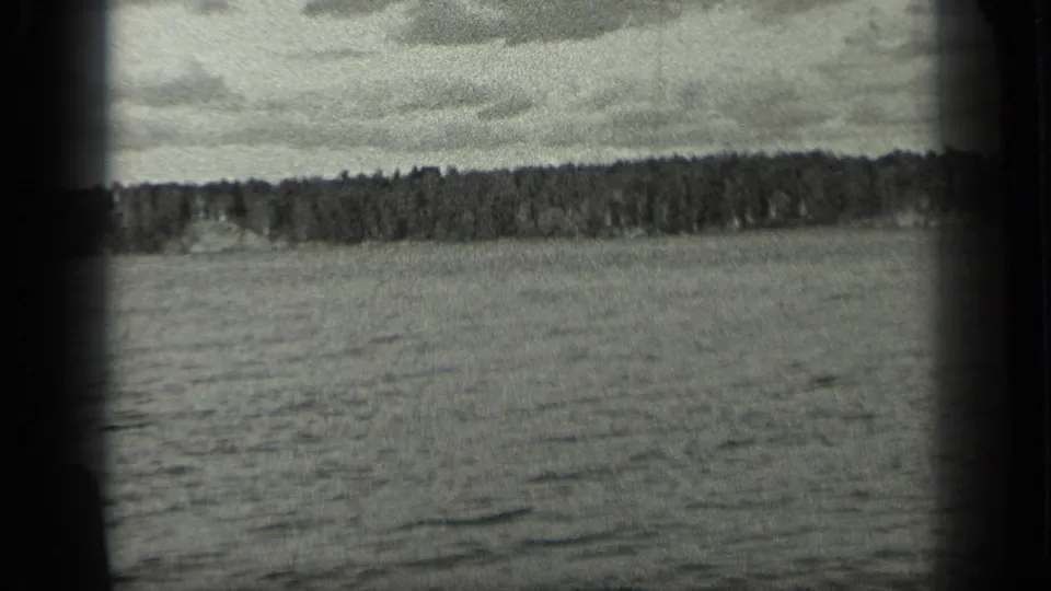 1939 Long Lake, Canada frame with open water in the foreground and a continuous wooded shoreline below a clouded sky.