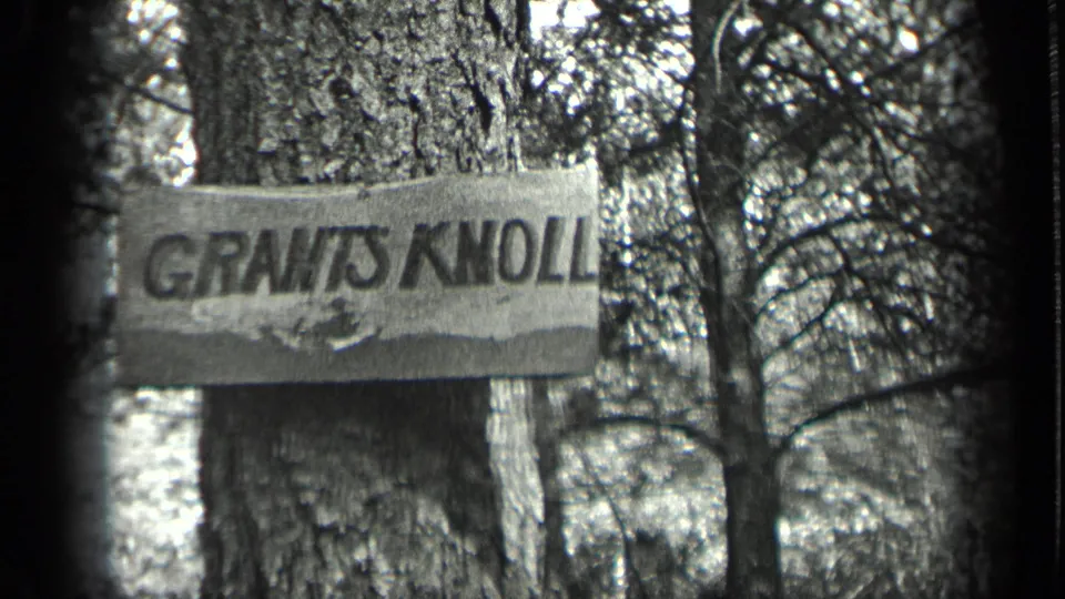 A GRANTS KNOLL sign is fixed to a tree trunk in the Long Lake forest, with branches and woods behind it.