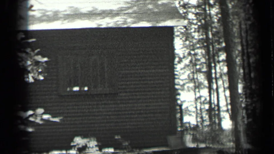 Long Lake, Canada cabin exterior in black-and-white 8mm, showing a dark wall, small windows, and trees around the structure.