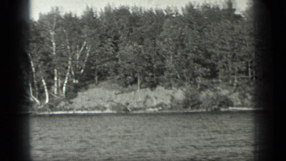Black-and-white 8mm view of Long Lake, Canada, showing water, a low bank, pale trunks, and dense shoreline trees.
