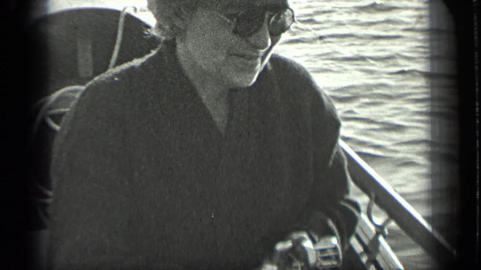 1939 Long Lake home-movie frame showing a woman in sunglasses seated on a boat, with fishing gear and rippling water nearby.