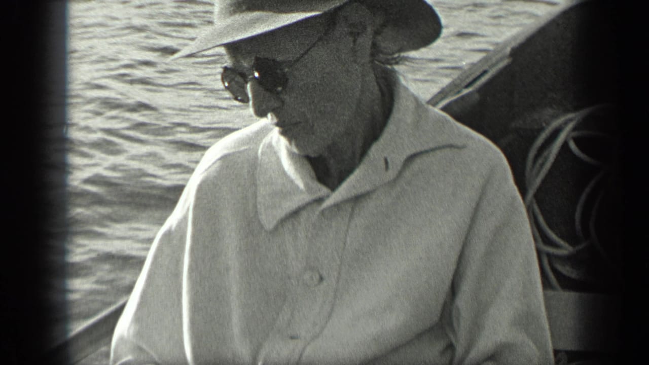 Man in sunglasses and a wide-brimmed hat sits on a boat near the water.