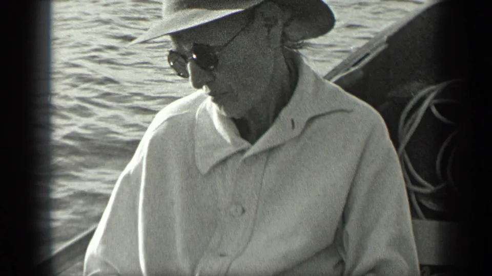 1939 Long Lake home-movie frame showing a man in a wide-brimmed hat and sunglasses seated on a boat beside rippling lake water.