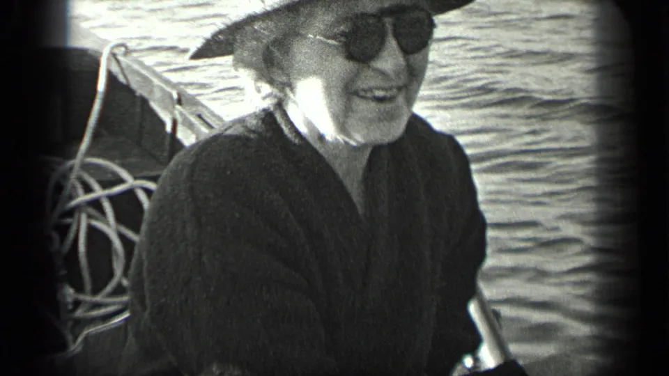 Older woman wearing dark sunglasses sits smiling in a small boat on Long Lake, Canada in 1939 home-movie footage.