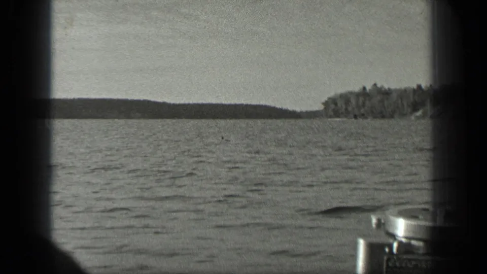 1939 Long Lake, Canada home-movie frame with open lake water, wooded shoreline, pale sky, and partial foreground hardware at the edge.