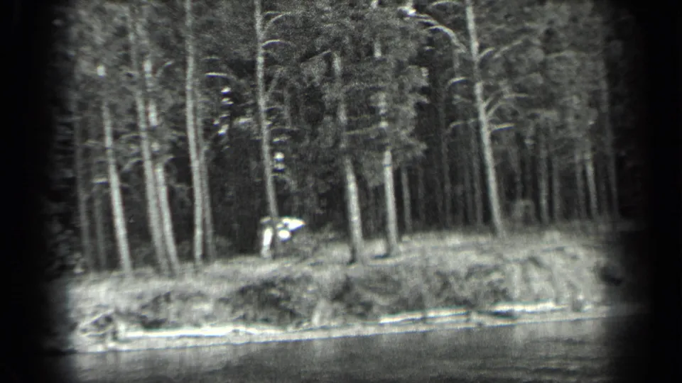 Black-and-white 8mm view of the Long Lake shoreline, showing forest, a low bank, and a distant light-toned figure or craft.