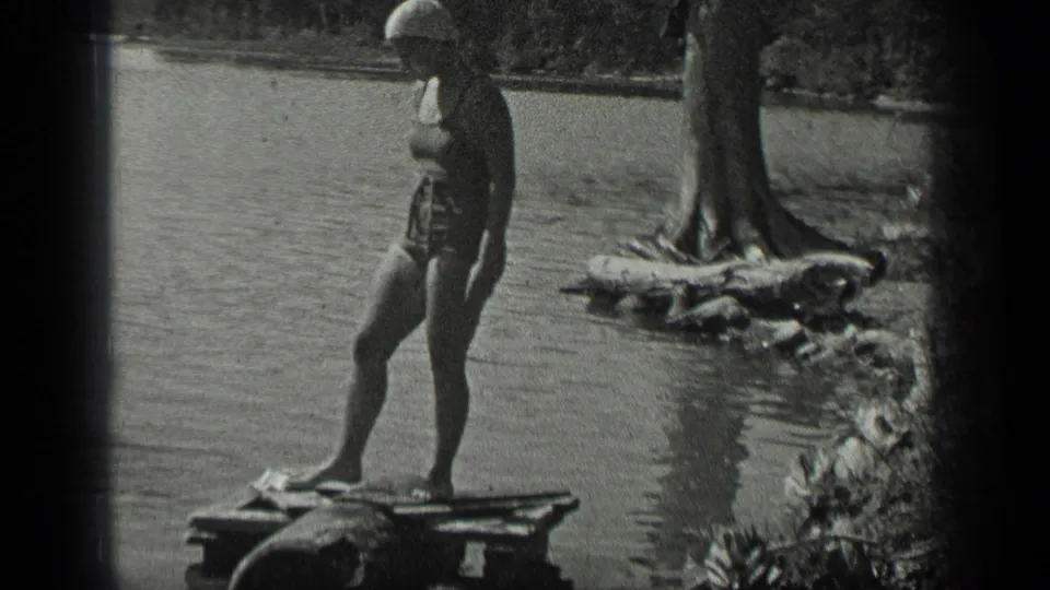1939 Long Lake frame showing a woman in a one-piece swimsuit and swim cap standing on a wooden platform at the shoreline.