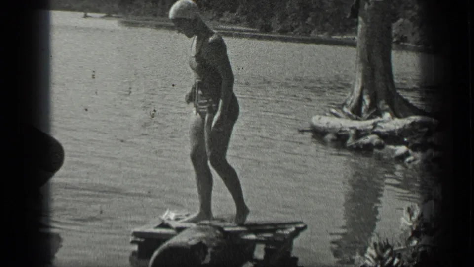 At Long Lake, Canada in 1939, a woman in a swim cap and swimsuit stands on a small wooden dock by the water.