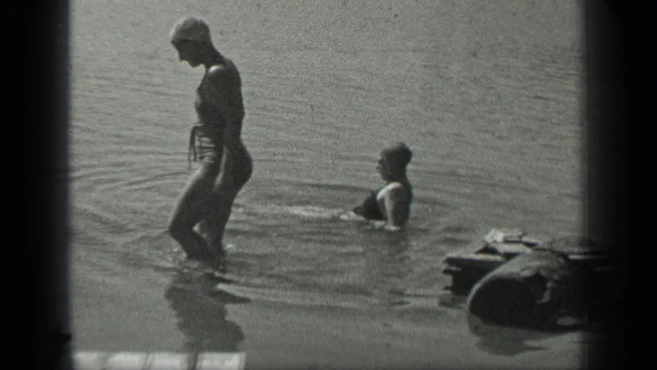 At Long Lake, Canada in 1939, a woman in a swim cap stands knee-deep as another swimmer rises from the lake.