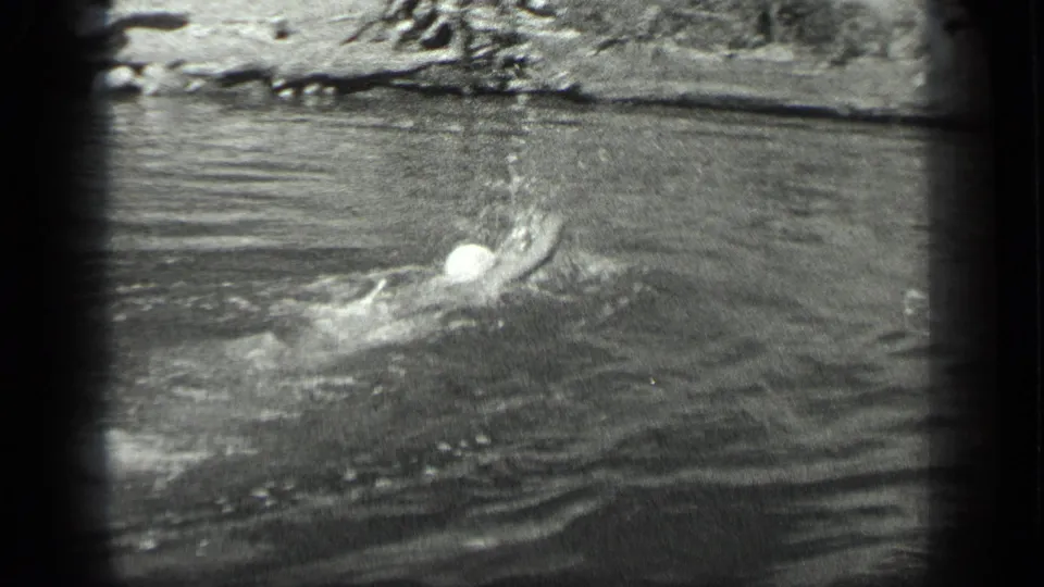 The swimmer appears mid-stroke in the Long Lake water, with a bright splash and shoreline visible behind.