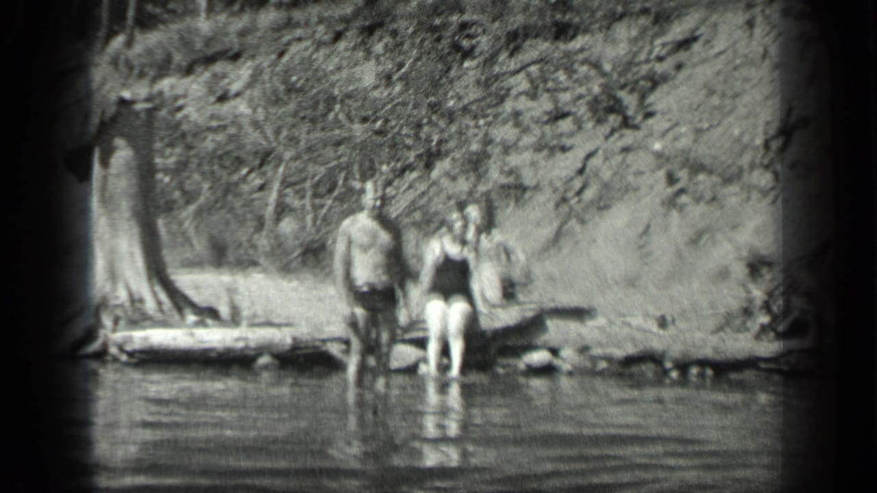 Three swimmers standing in shallow lake water beside a wooded shoreline.
