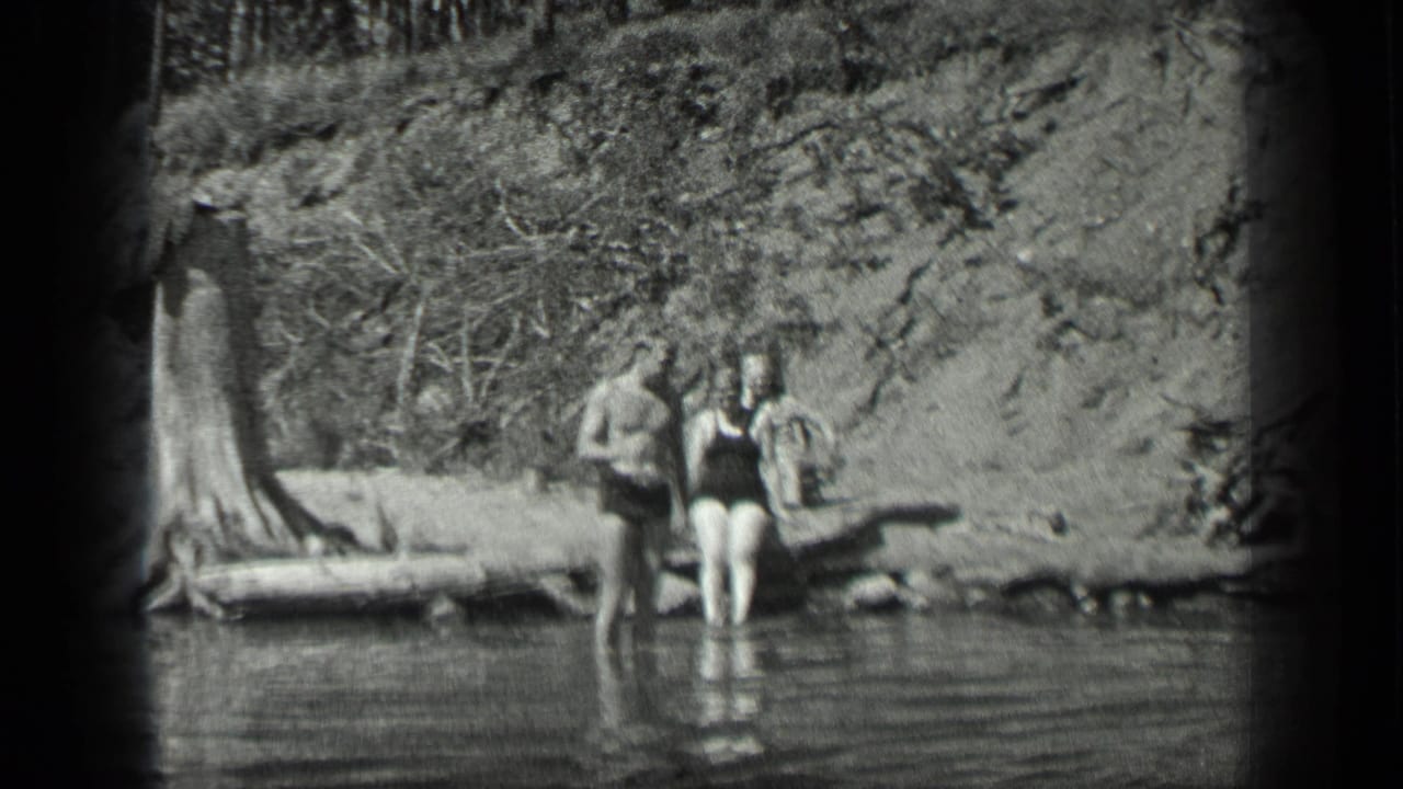 Home-movie view of three swimmers at Long Lake, Canada, with a wooded bank and still water in the foreground.