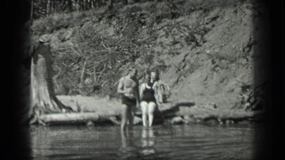 Home-movie view of three swimmers at Long Lake, Canada, with a wooded bank and still water in the foreground.