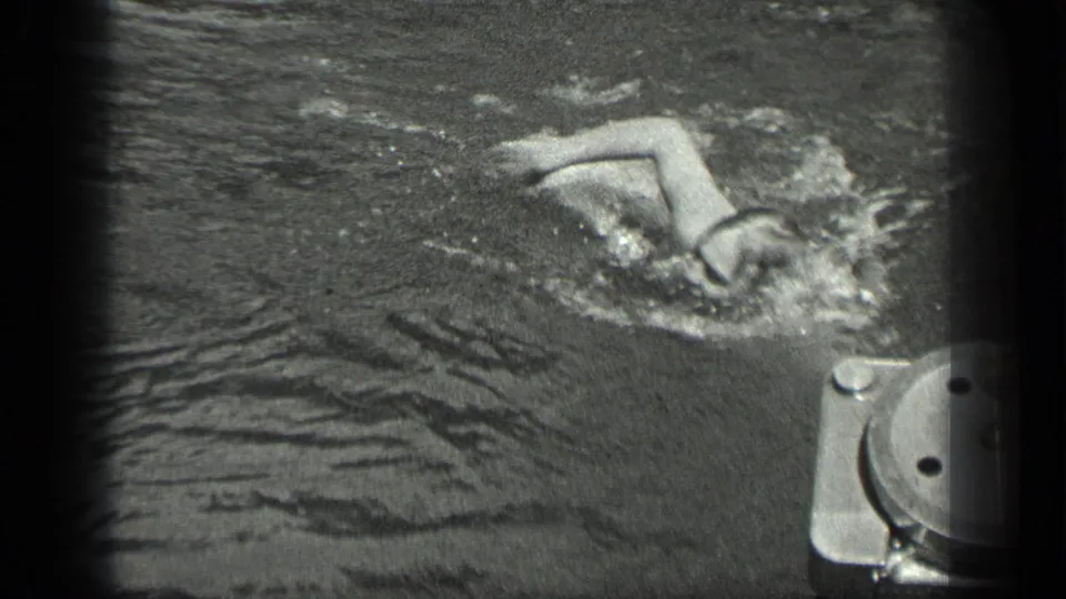 A swimmer in dark water raises an arm during a stroke near dock or boat hardware at Long Lake, Canada.