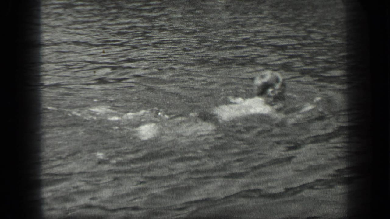 Swimmer moving through rippled lake water in black-and-white footage.