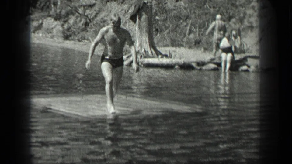 A person in swimwear steps from a lakeside dock or platform as other bathers stand near the wooded shore at Long Lake.