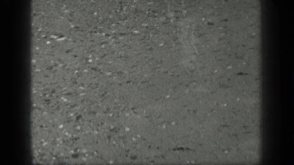1939 Long Lake, Canada 8mm frame showing rain-darkened concrete or pavement with scattered grit and heavy edge shadowing.