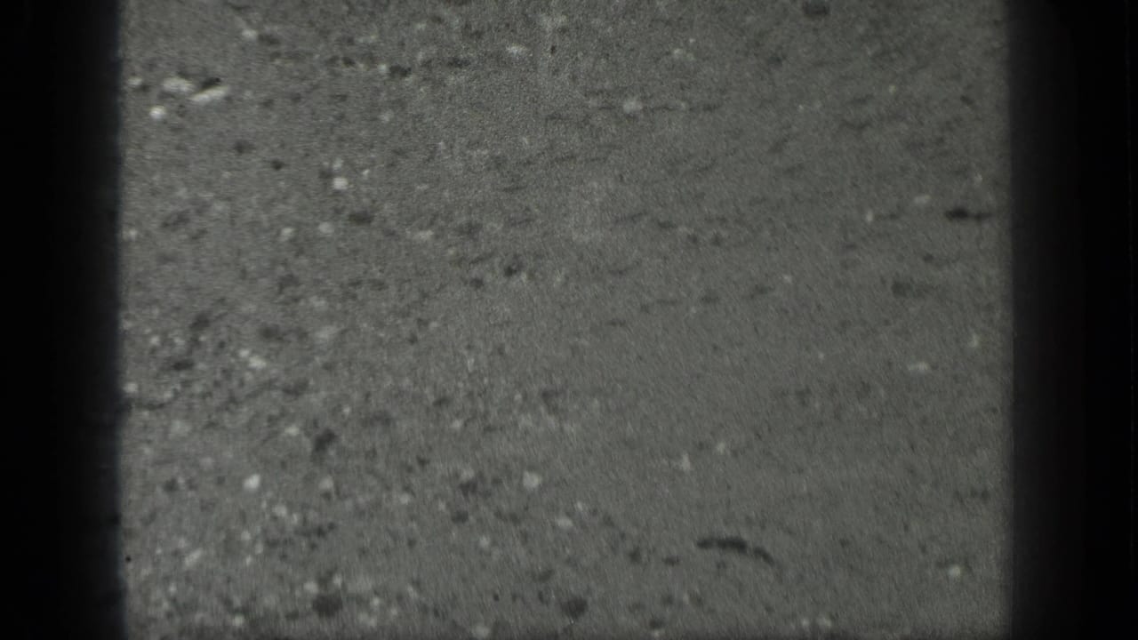 1939 Long Lake, Canada 8mm frame of wet pavement or concrete under rain, with grain, scratches, and dark vertical gate shadow.