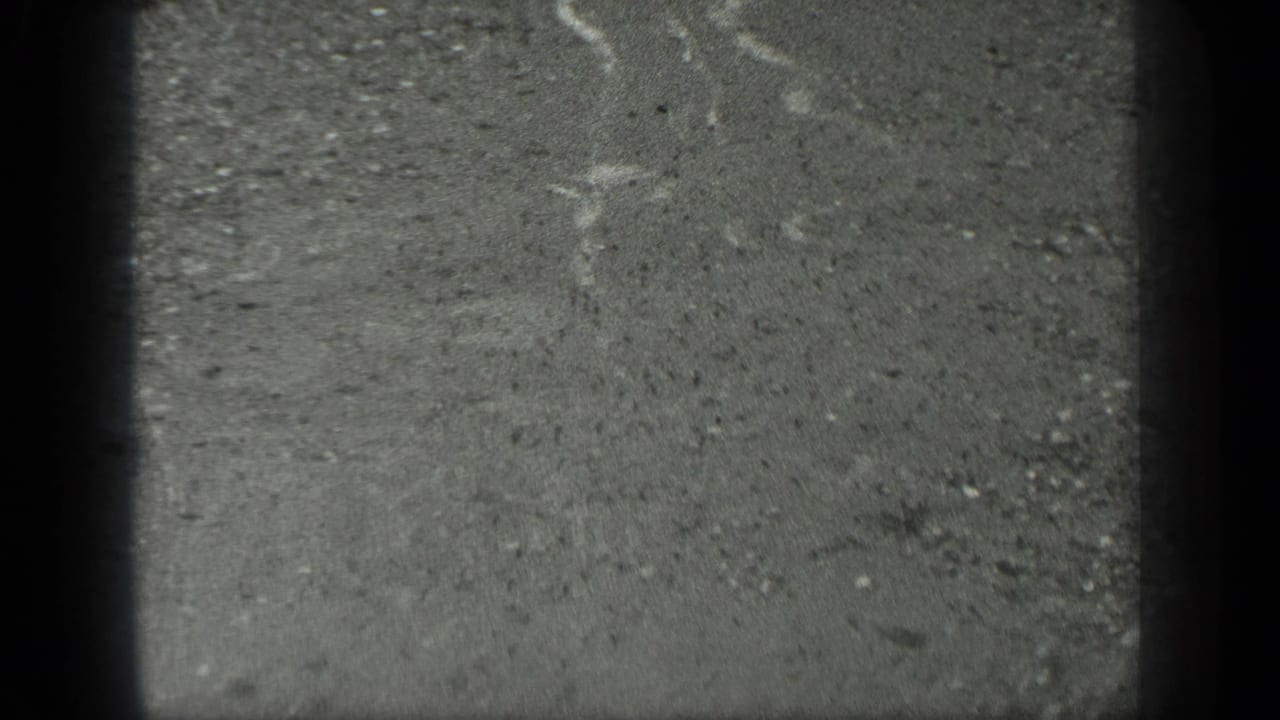 1939 Long Lake, Canada 8mm frame of rain-wet concrete or pavement with pale water streaks, scattered grit, and dark edge shadowing.