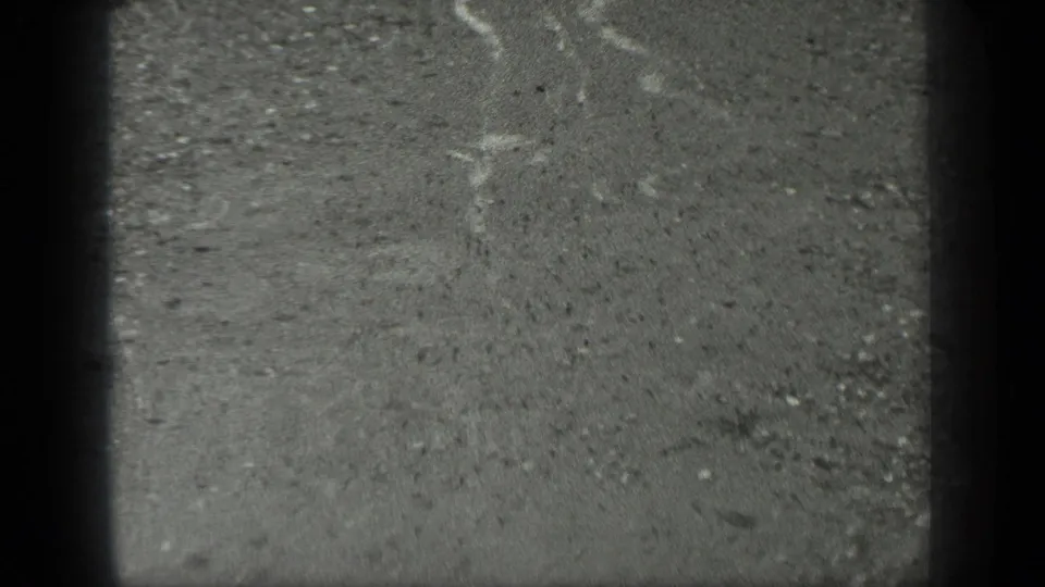1939 Long Lake, Canada 8mm frame of rain-wet concrete or pavement with pale water streaks, scattered grit, and dark edge shadowing.