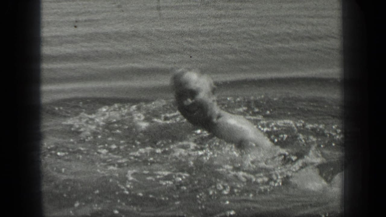 A man swims in Long Lake, Canada, in black-and-white 8mm footage, his head and shoulder briefly visible above splashing water.