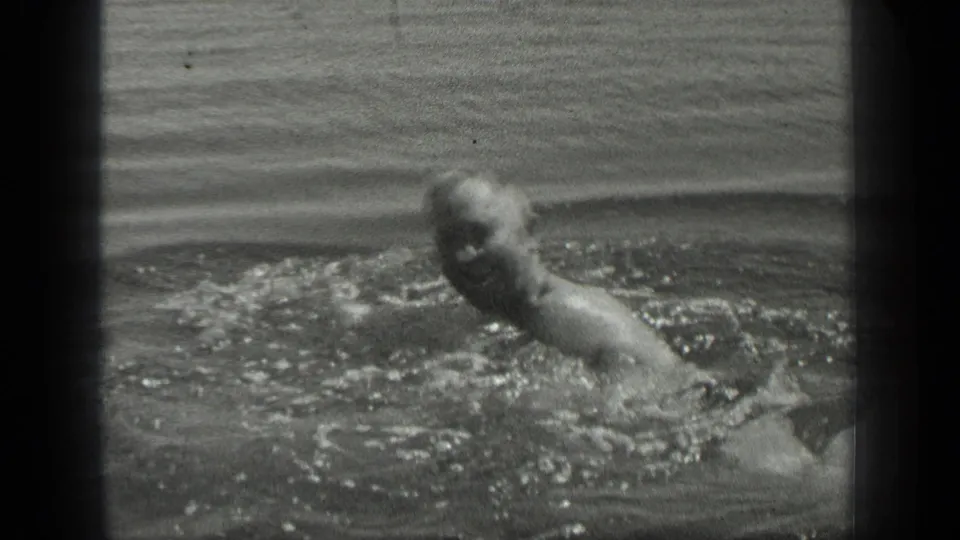 A man swims in Long Lake, Canada, in black-and-white 8mm footage, his head and shoulder briefly visible above splashing water.