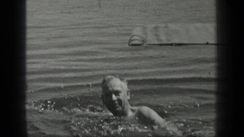 The adult swimmer moves toward the camera at Long Lake, Canada, with splashing water and a wooden pier in the background.