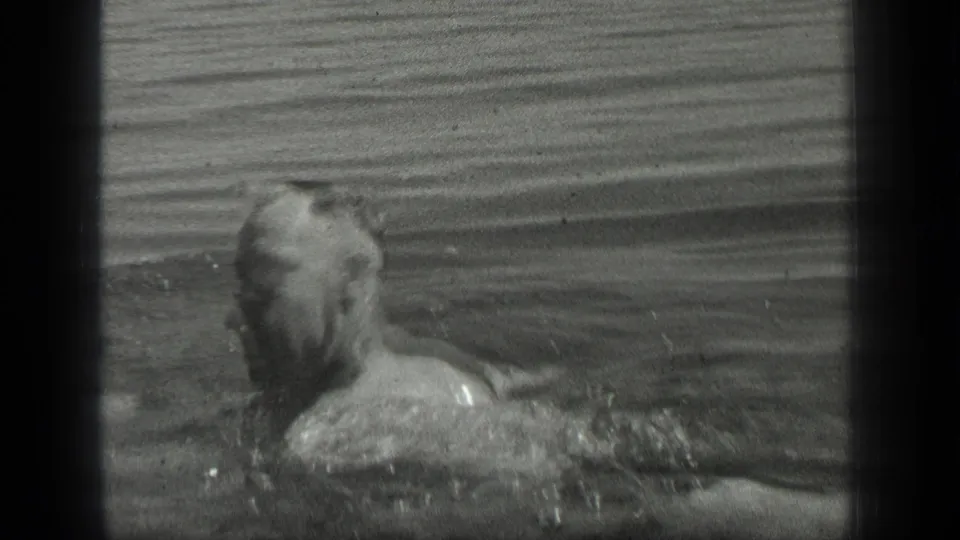 The swimmer turns his head and shoulders sideways in the lake, with 8mm grain and black frame edges visible.