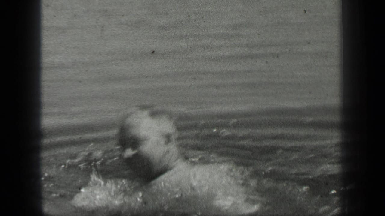 An adult swimmer moves across the frame in black-and-white 8mm footage, with water splashing near his arm and face partly visible.