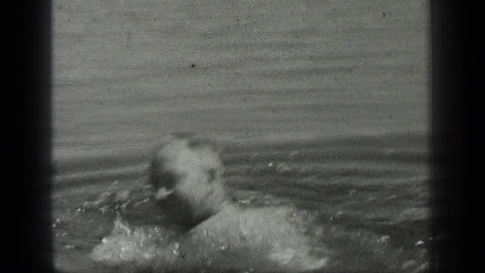 An adult swimmer moves across the frame in black-and-white 8mm footage, with water splashing near his arm and face partly visible.