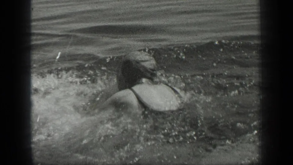 In 1939 Long Lake, Canada footage, a woman in dark swimwear swims through rippling lake water, seen from behind.