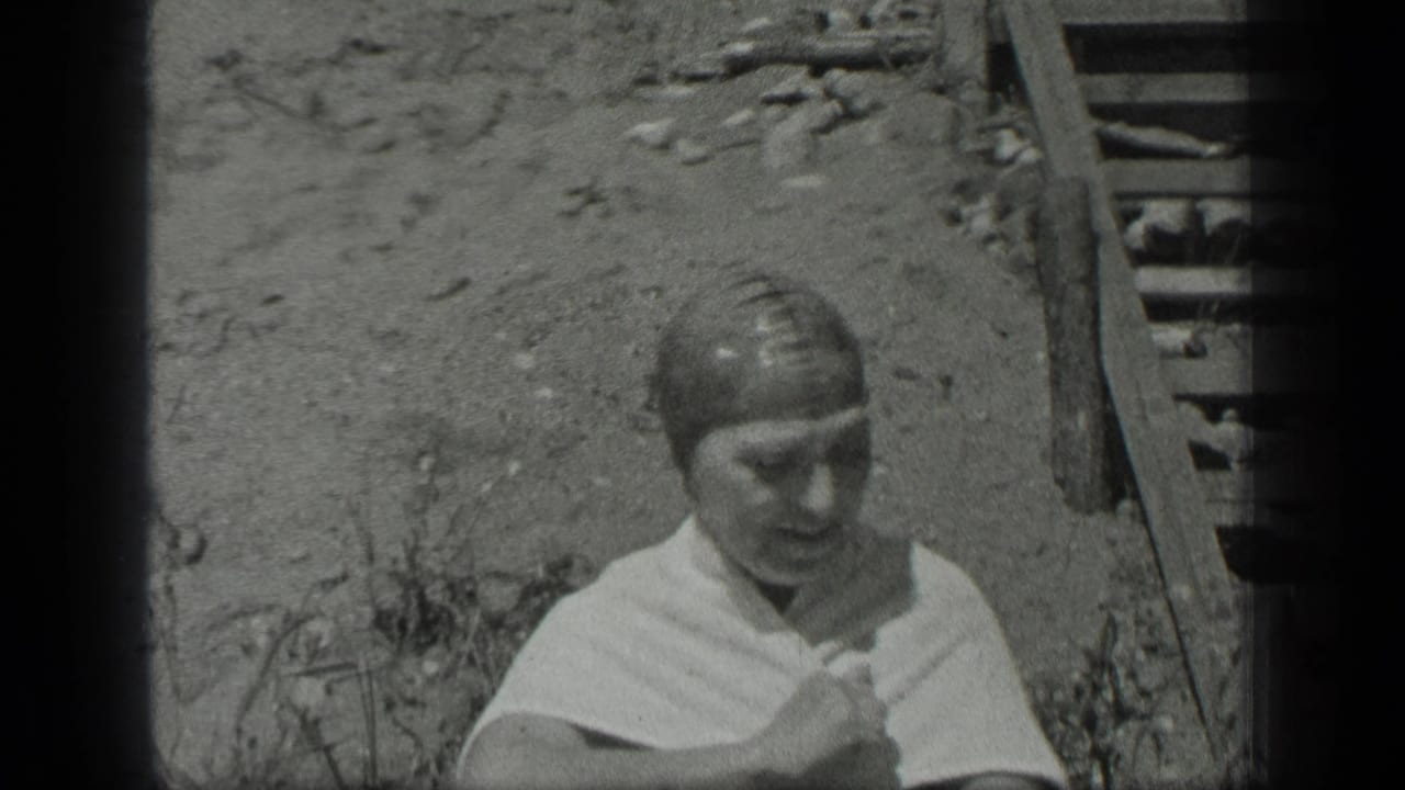 Woman wrapped in a towel seated near a wooden ladder on rough beach ground.