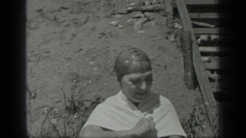 At Long Lake, Canada, a towel-wrapped woman sits on a rough shoreline near a simple wooden ladder in 1939 home-movie footage.