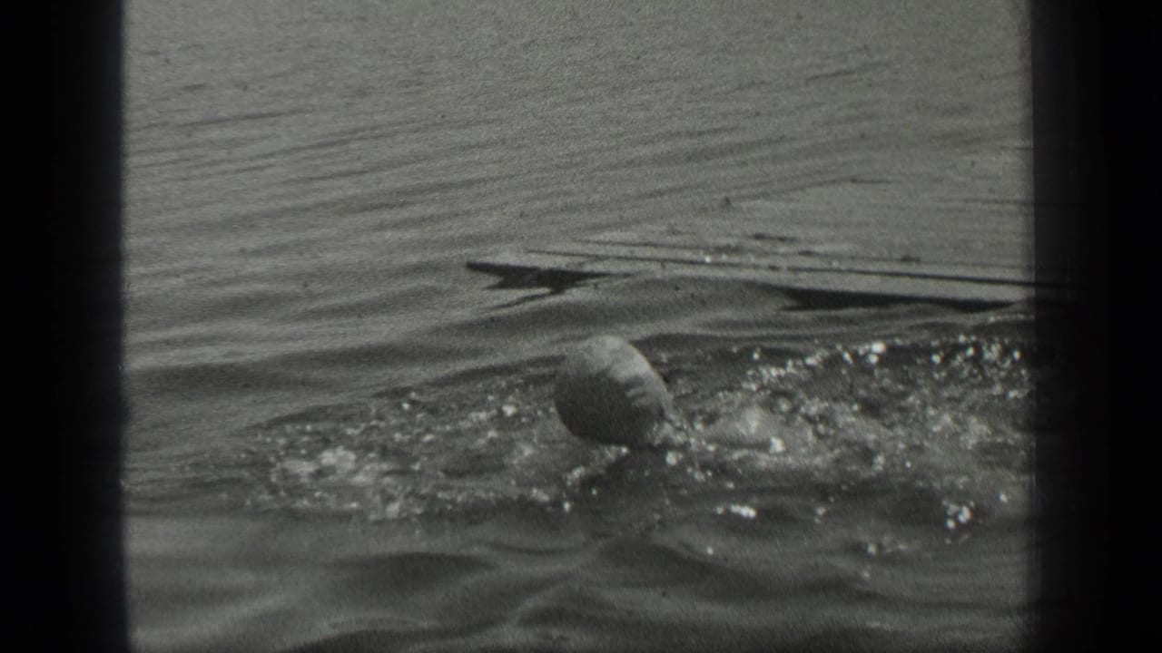 A woman swims in rippled Long Lake water, with her head and arm breaking the surface in black-and-white 8mm footage.