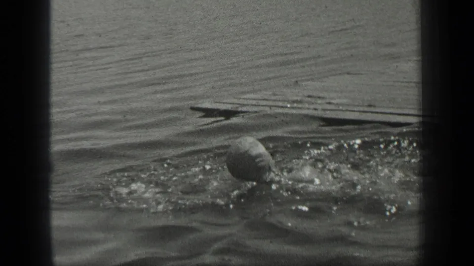 A woman swims in rippled Long Lake water, with her head and arm breaking the surface in black-and-white 8mm footage.