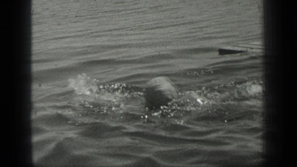 The woman continues swimming across calm Long Lake water, her head and arm partly visible among ripples and reflected light.