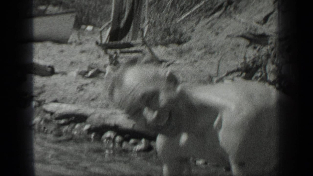 1939 Long Lake, Canada frame showing a man reclining or leaning near the rocky lakeshore, with a wooden chair visible behind him.
